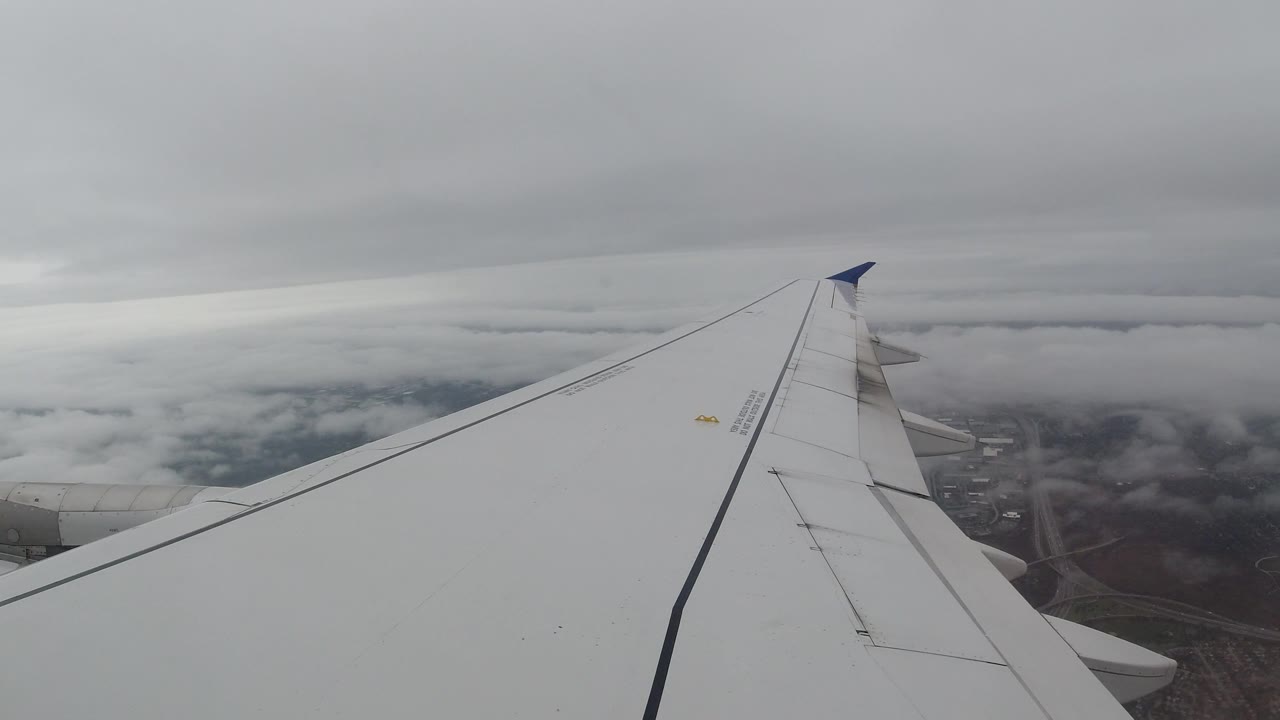 A white airplane wing extends across a cloudy sky over Los Cabos, Mexico, with muted land and roads barely visible below and no visible people.