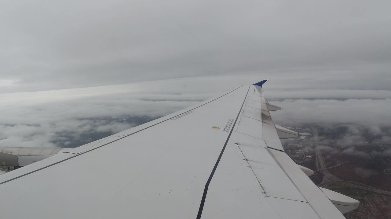 The airplane wing remains centered beneath a thick overcast layer over Los Cabos, Mexico, with patchy terrain showing through the clouds and no visible people.
