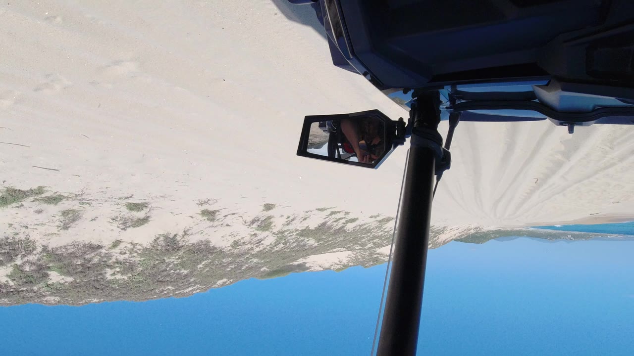 Upside-down vehicle view over sand, scrub, ocean, and blue sky.