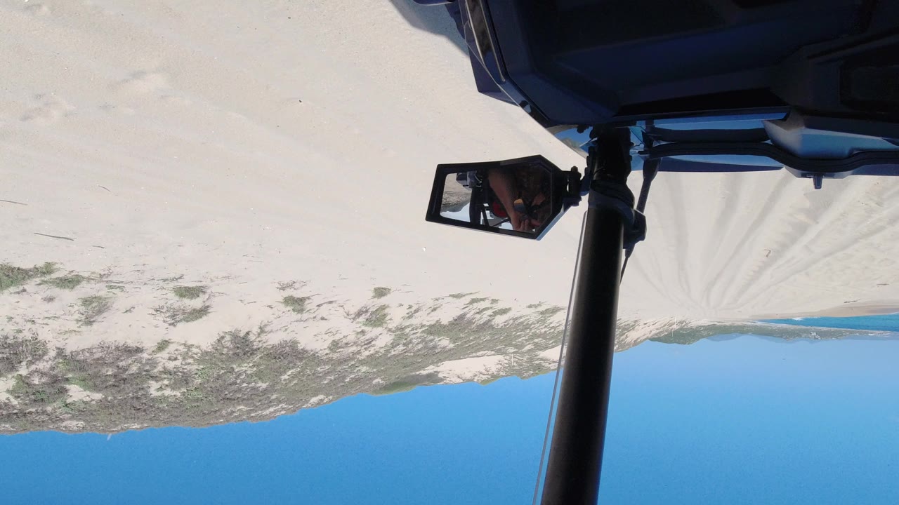 A later upside-down view in Los Cabos, Mexico, keeps the vehicle roll bar and mirror in front while the sandy beach, scrub line, ocean, and clear sky fill the background with no visible people.