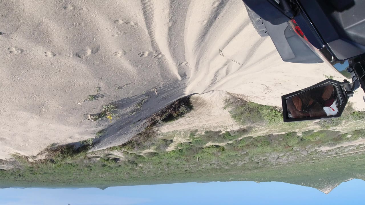 Upside-down view in Los Cabos, Mexico, showing pale sand, scattered shrubs, distant hills, blue sky, and dark camera or gear along the upper right edge, with no visible people.