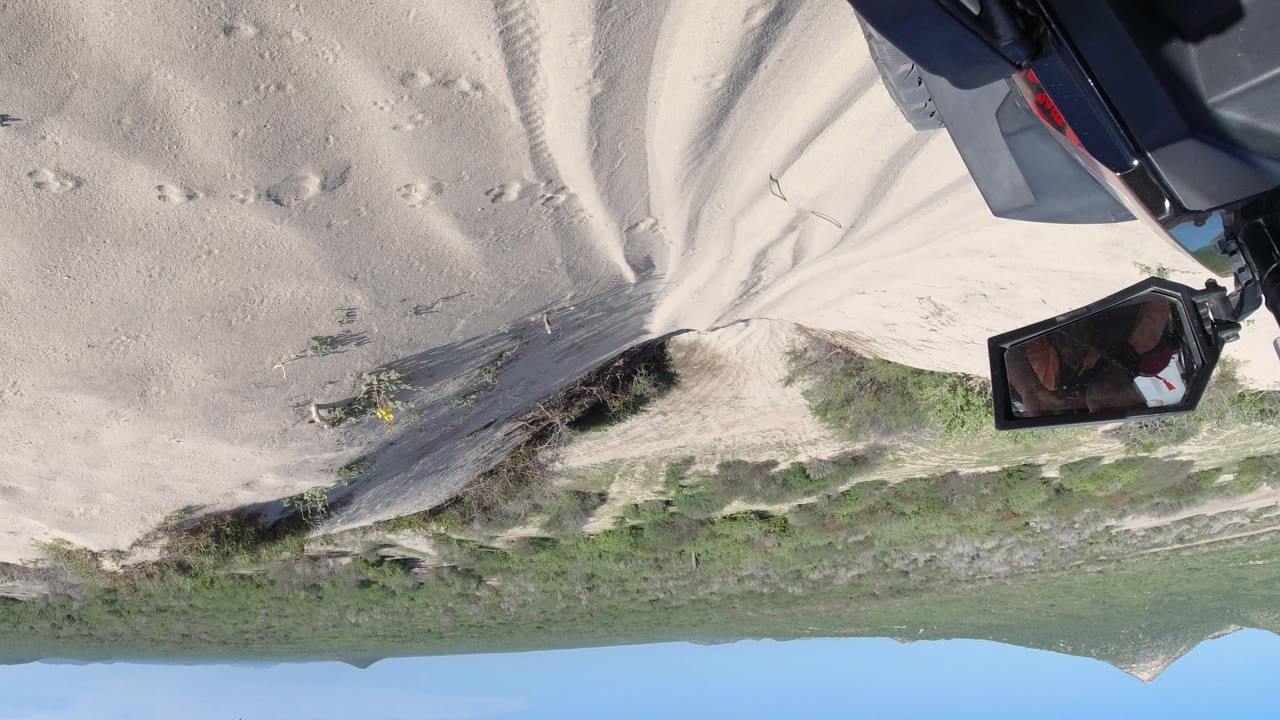 Upside-down Los Cabos hillside frame with sand tracks, scrub vegetation, a mountain ridge, clear blue sky, and a dark piece of gear still visible near the upper right, with no visible people.