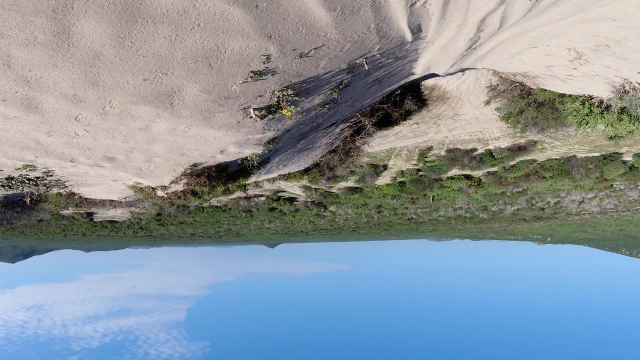 Upside-down aerial frame of a pale sandy slope in Los Cabos, Mexico, with sparse green scrub, a distant horizon, blue sky along the bottom, and no visible people.