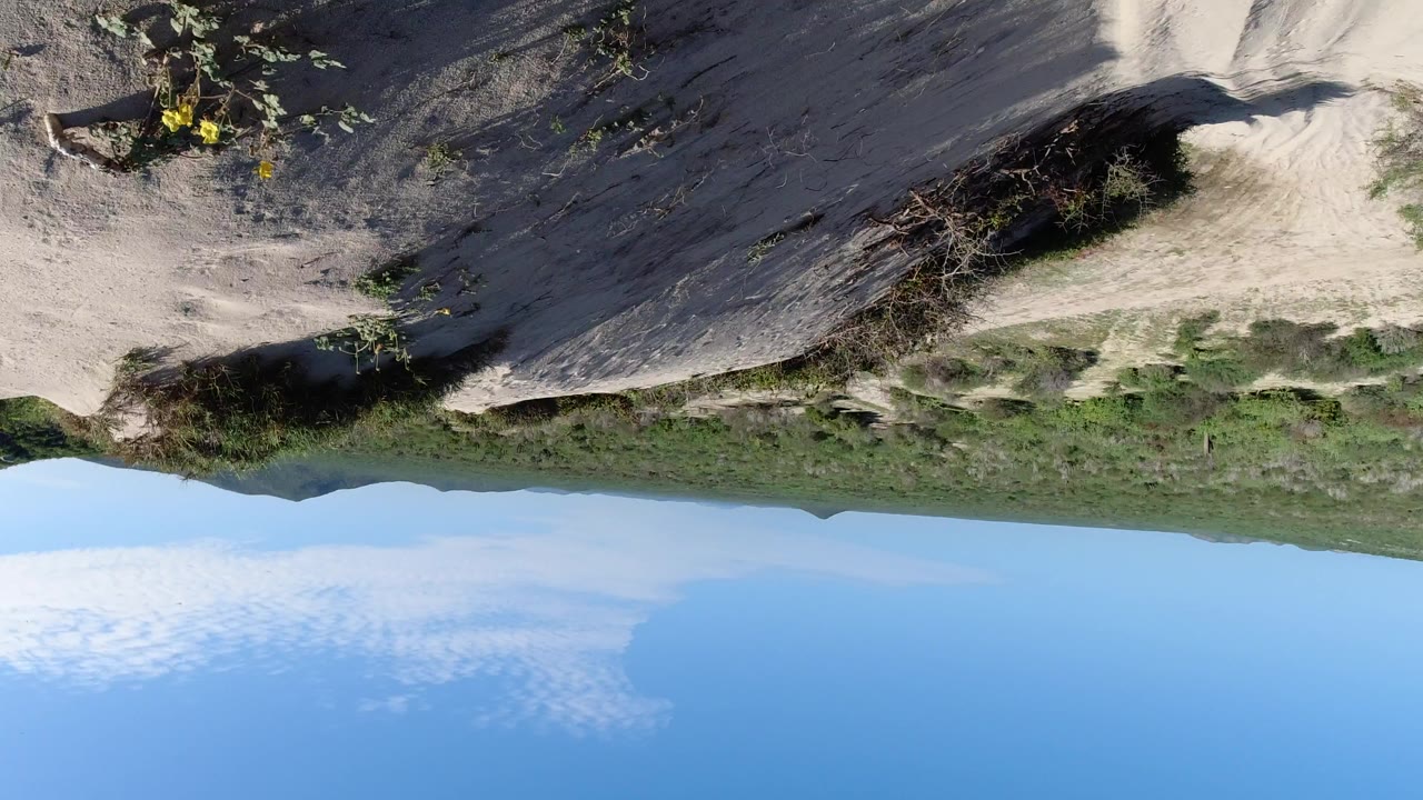 Upside-down aerial view in Los Cabos, Mexico, showing pale sandy hillside, scattered brush, distant green ridge, blue sky, and no visible people.