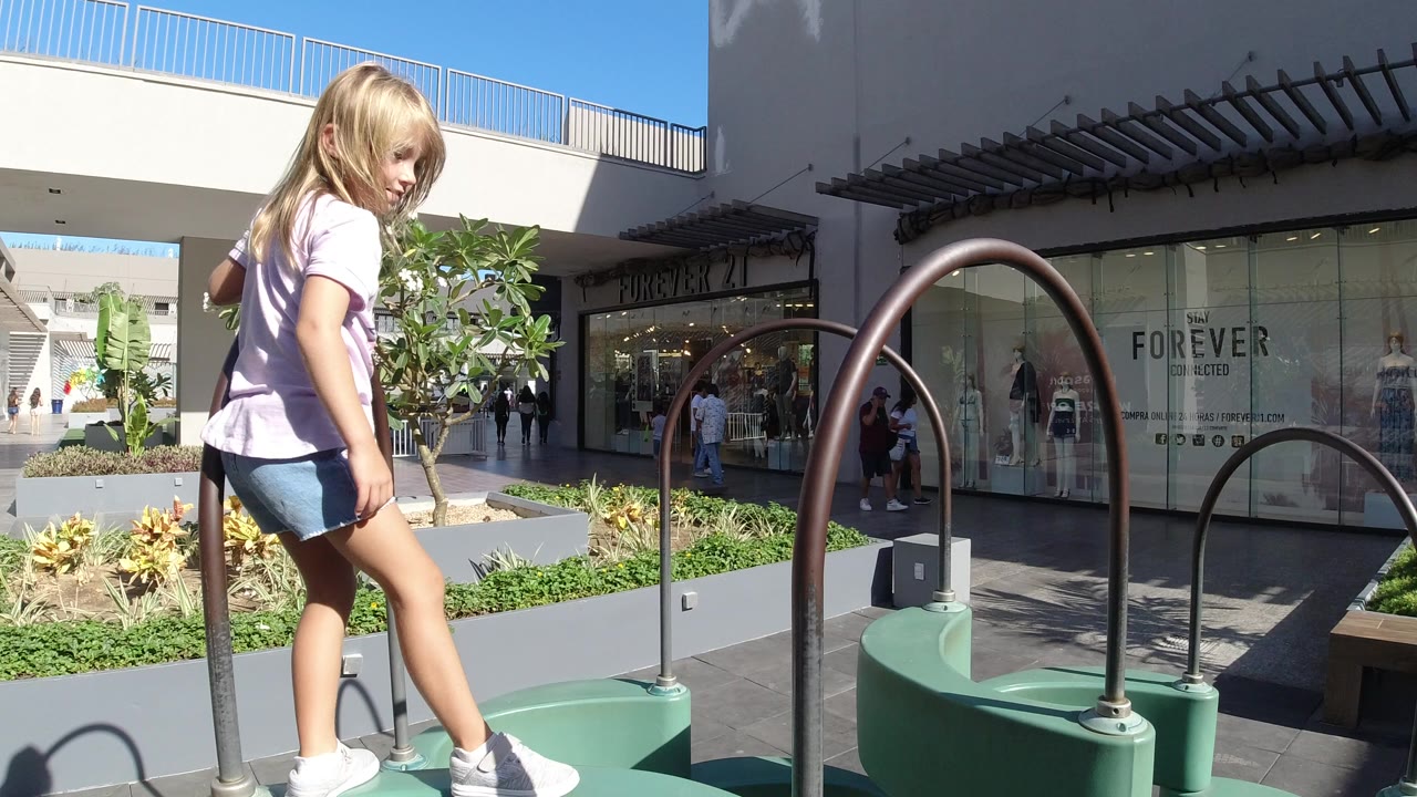 A young child stands on green play equipment beside curved metal rails in a Los Cabos shopping plaza, with storefronts, planters, and pedestrians behind her.