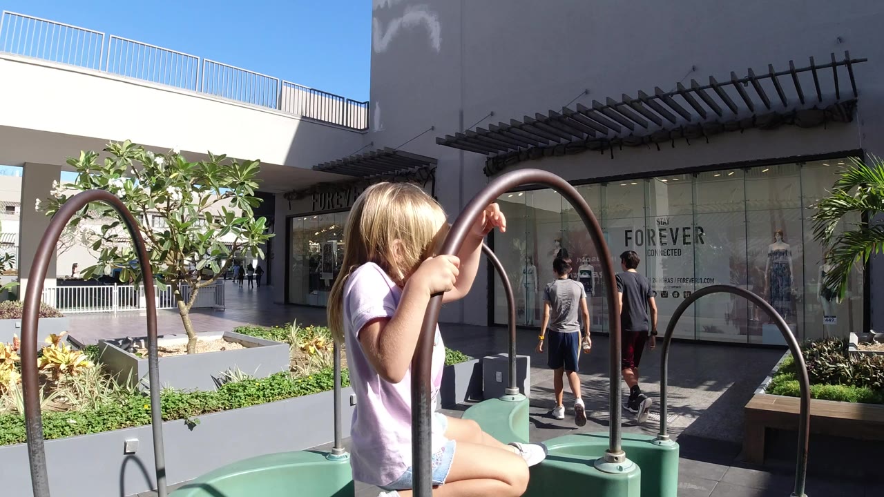 The girl holds a tall brown hoop on the green obstacle toy in Los Cabos, Mexico, while two pedestrians walk near glass storefronts in the background.