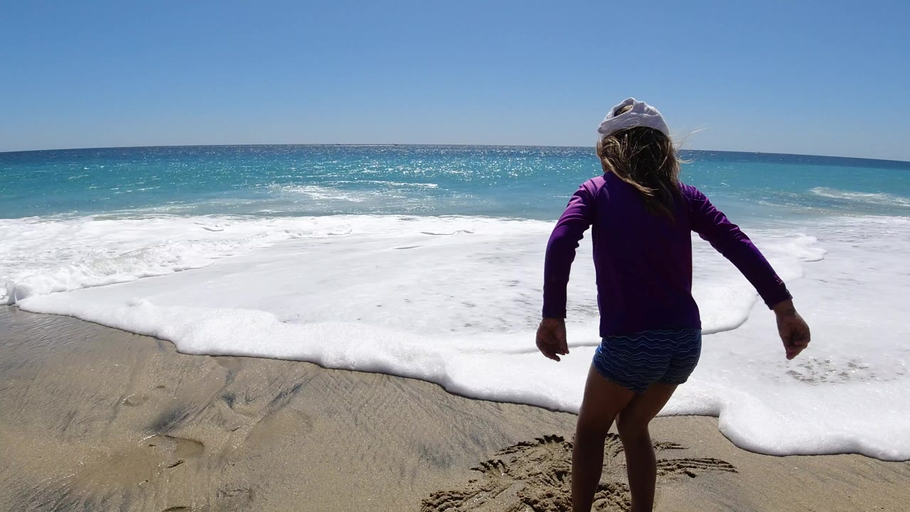 Girl in white hat facing ocean waves on sandy beach.