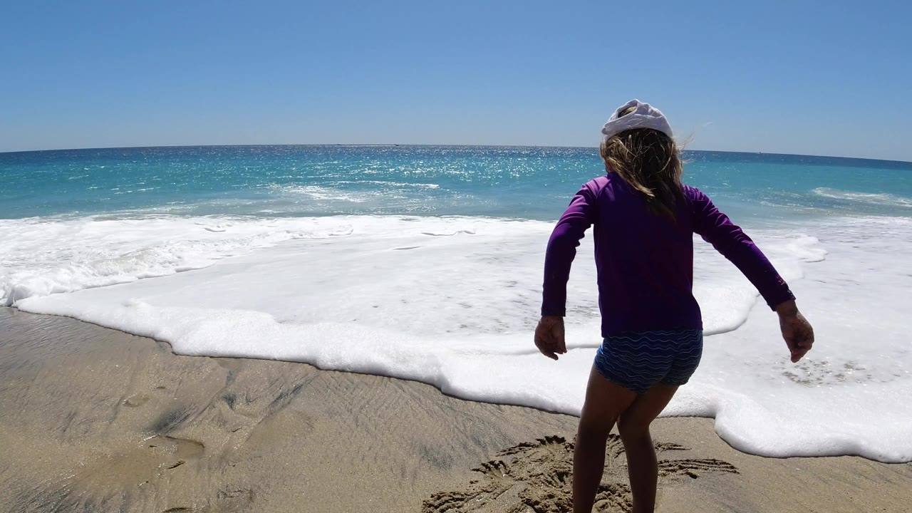 The girl steps closer to the white wash on the Los Cabos shoreline, with wet sand in the foreground and turquoise water behind her.