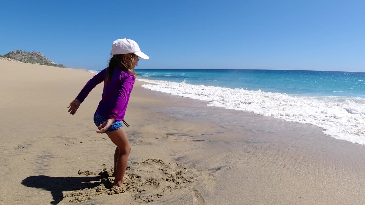 Child in a white cap stands on wet sand beside incoming surf.