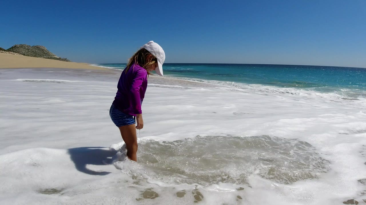 Person in a white cap standing in foamy surf on a sandy beach.