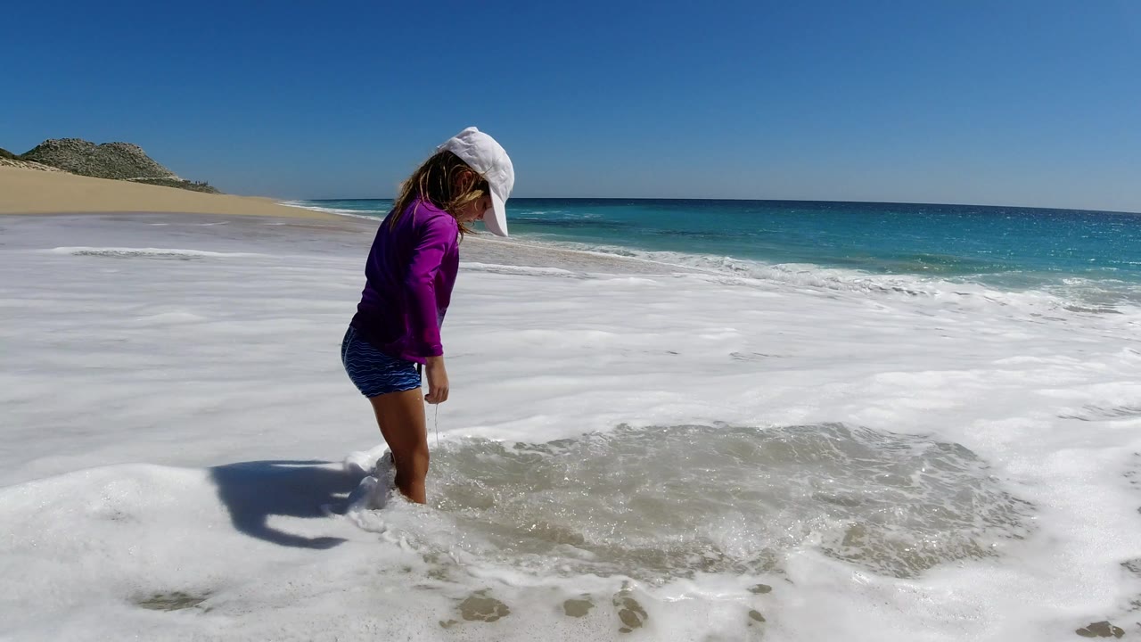 At 0:01 in Los Cabos, Mexico, the person remains near the shoreline while a sheet of white foam pushes around their calves under strong daylight.