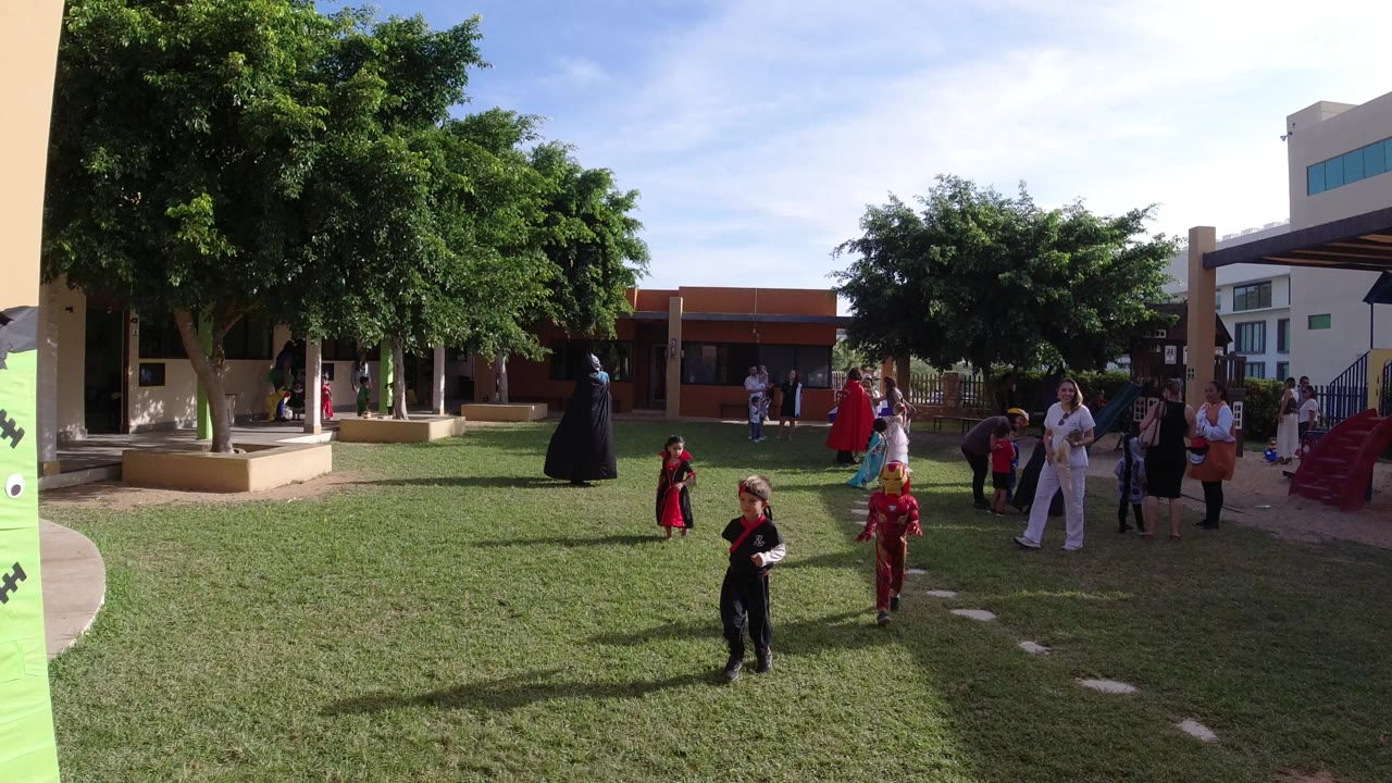 Wide view in Los Cabos, Mexico, showing children in costumes and adults standing around a sunny grass courtyard with trees, buildings, and playground equipment.