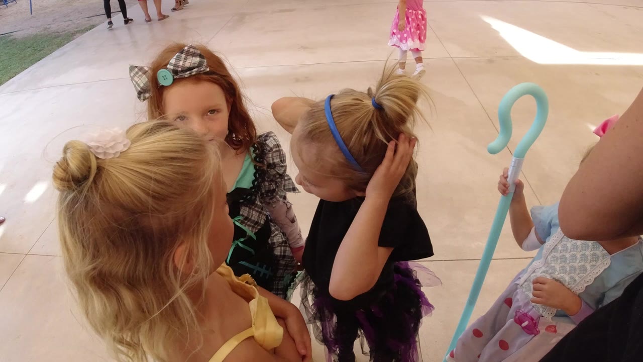 Young girls in costumes cluster on a sunlit tiled patio in Los Cabos, Mexico, with a blue hook prop and adults partly visible at the edge of the frame.