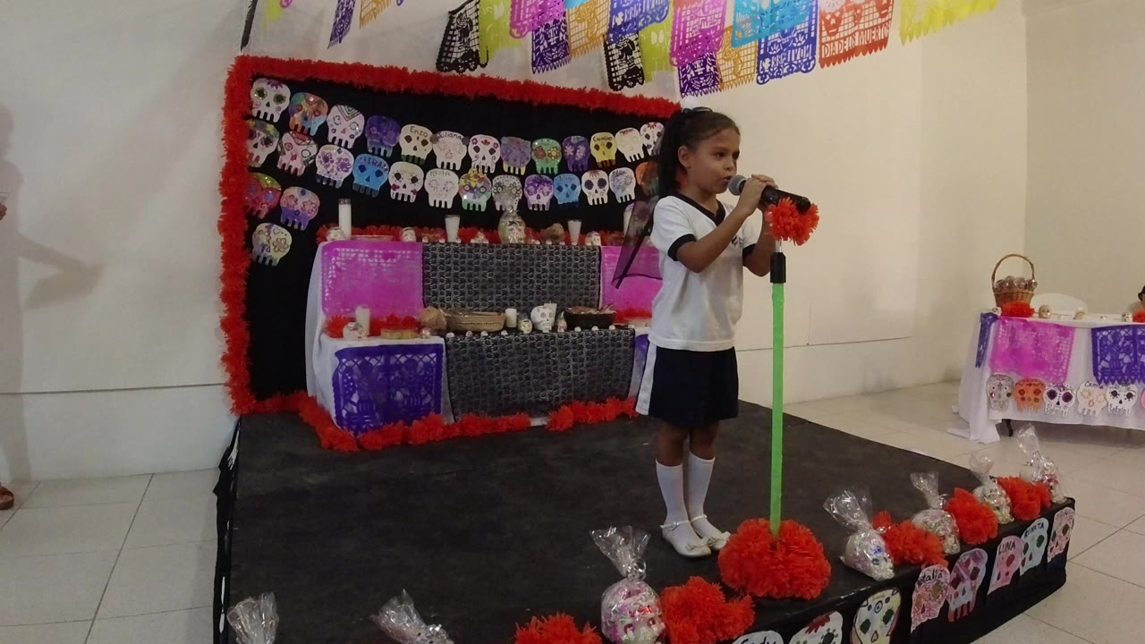 At 0:00, a child in a school uniform stands at a green microphone on a small decorated stage in Los Cabos, Mexico, with skull cutouts and papel picado behind her.