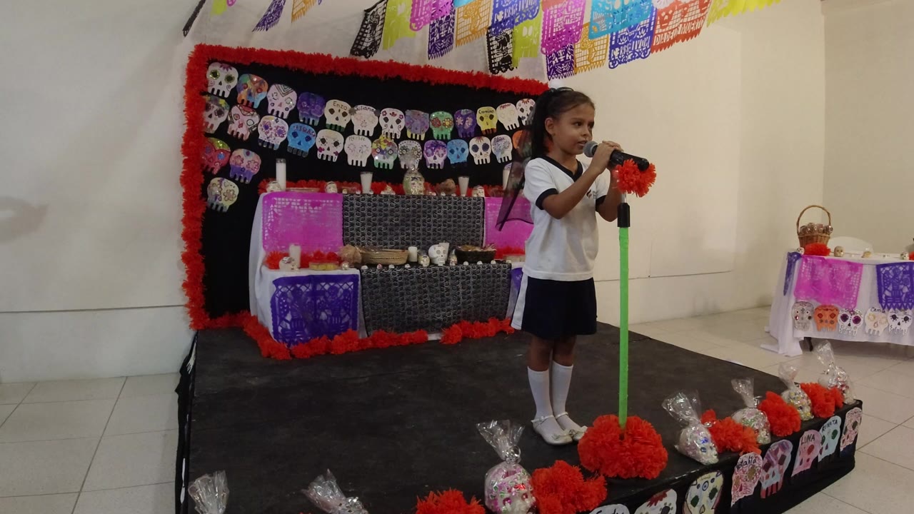 At 0:00 in Los Cabos, Mexico, a young girl stands at a green microphone stand on an indoor stage with skull artwork, papel picado banners, and red flower decorations behind her.
