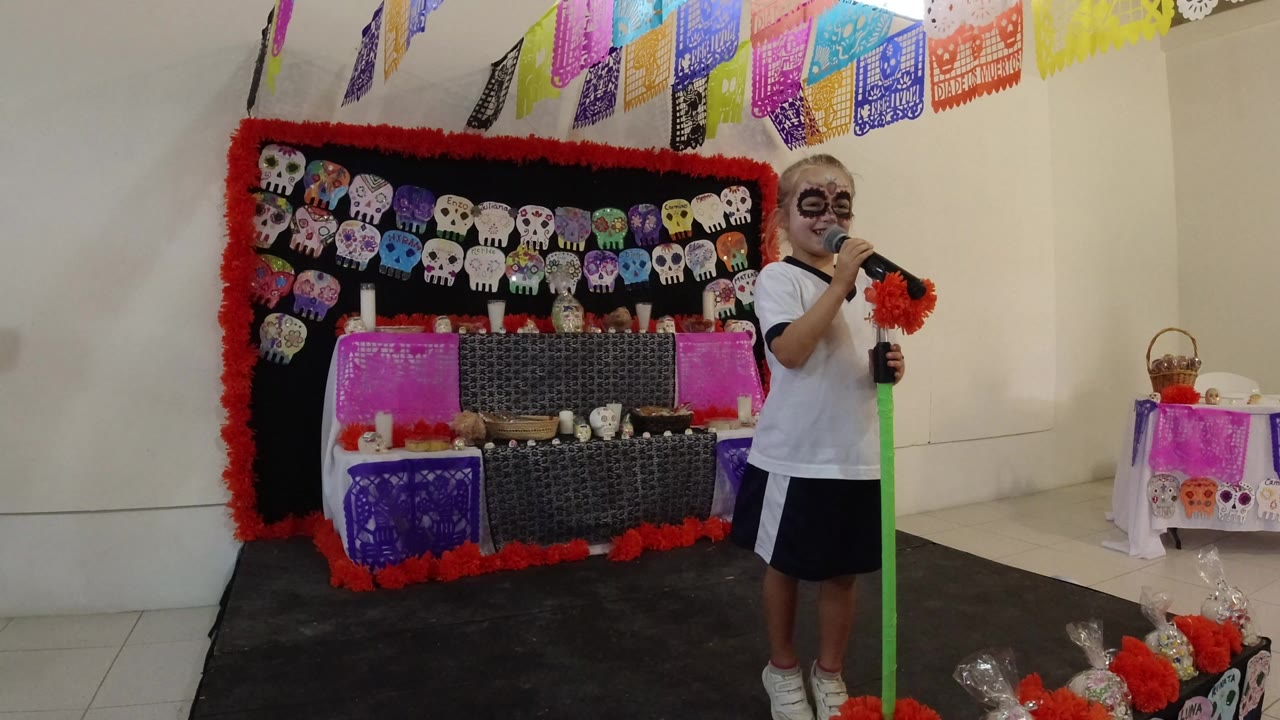 A young girl with skull-style face paint sings into a microphone on a small decorated stage in Los Cabos, Mexico, with colorful paper banners and skull artwork behind her.