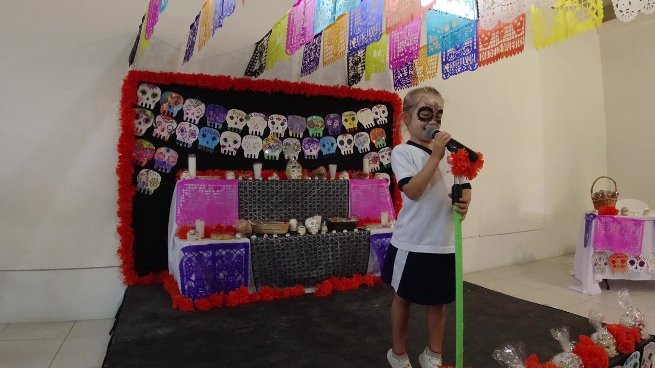 At 0:00 in Los Cabos, Mexico, a child with skull-style face paint stands near a green microphone stand in front of papel picado and a skull-decorated display.