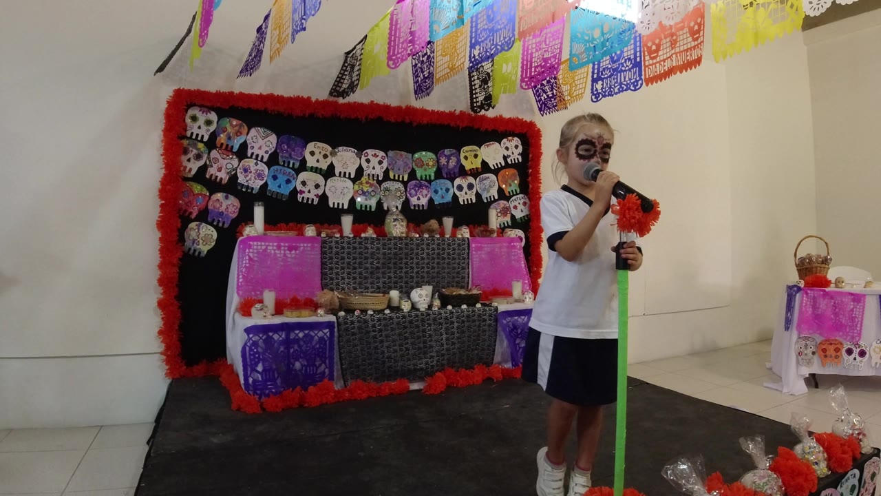 At 0:01, the girl remains near the right edge of the frame while the skull-decorated backdrop and overhead paper banners fill the stage area in Los Cabos, Mexico.