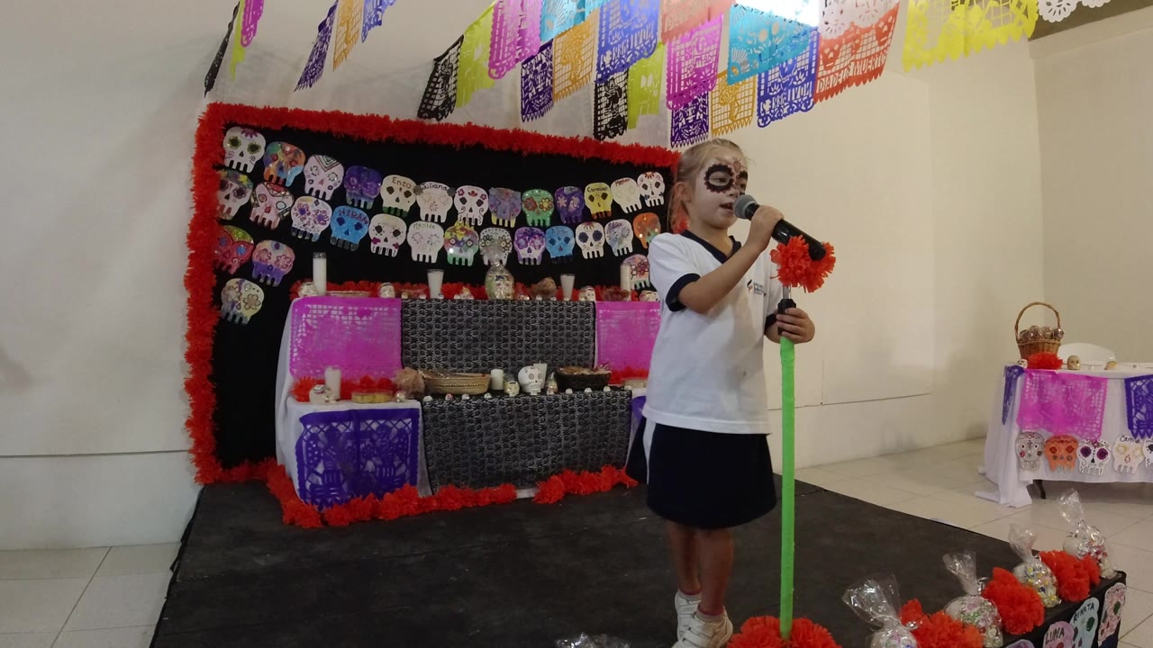 One second in, the child holds the microphone near their mouth while standing to the right of a skull-decorated backdrop and hanging papel picado in Los Cabos, Mexico.