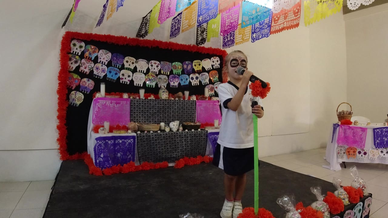 Child singing beside a Day of the Dead display with skull decorations.