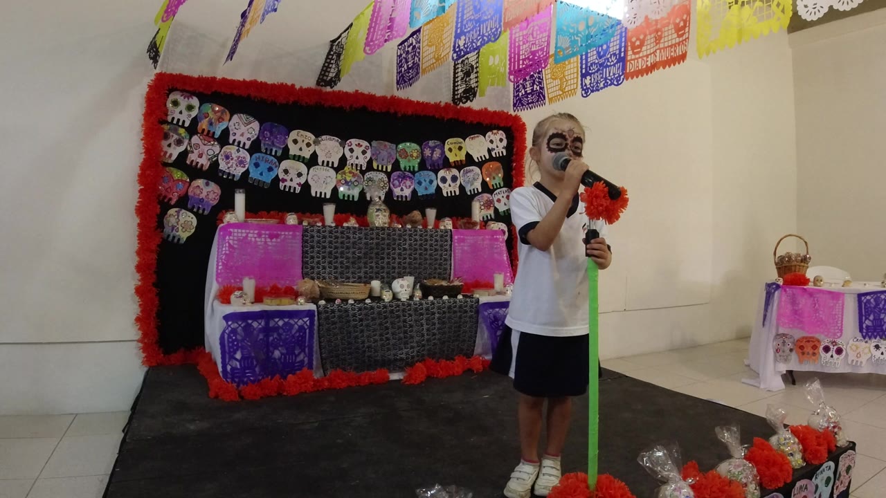 At 0:00 in Los Cabos, Mexico, a child with skull face paint stands near a green microphone stand beside a stage decorated with skull artwork, red garlands, and colorful paper banners.
