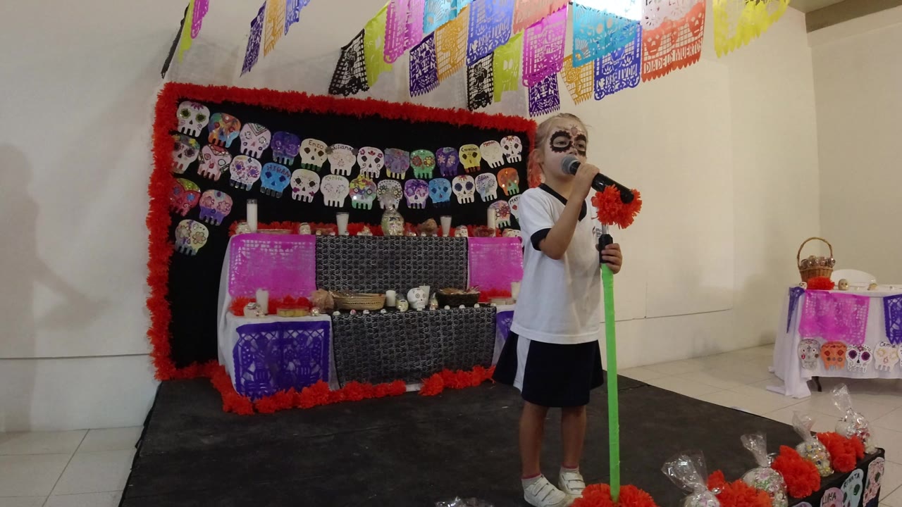 At 0:00, a child in Los Cabos, Mexico wears skull-style face paint while singing into a microphone beside a decorated skull backdrop and colorful paper banners.
