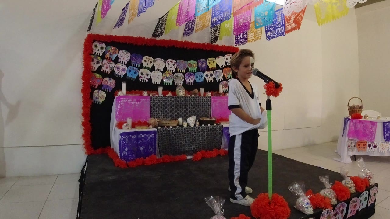A young boy in Los Cabos, Mexico stands near a green microphone on a small stage with papel picado, skull decorations, and red paper flowers behind him at the start of the clip.