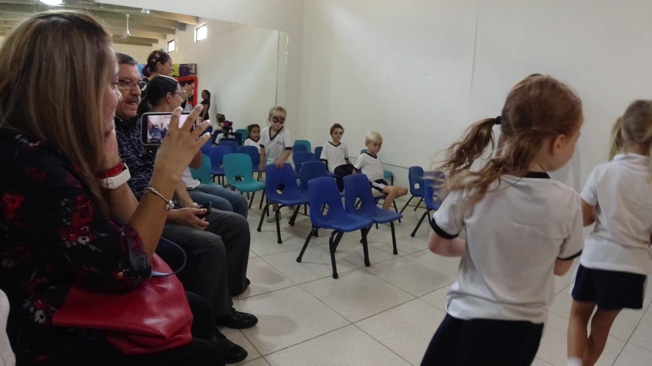 Parents in Los Cabos, Mexico sit along the side of a bright classroom while children in white uniforms move between rows of blue chairs.
