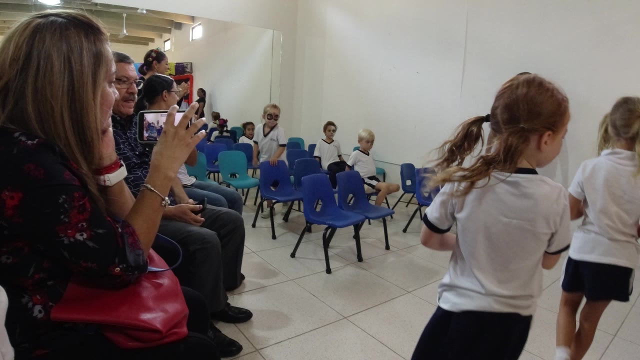 Two girls in white school uniforms stand near rows of blue chairs as adults watch and record inside the Los Cabos classroom.