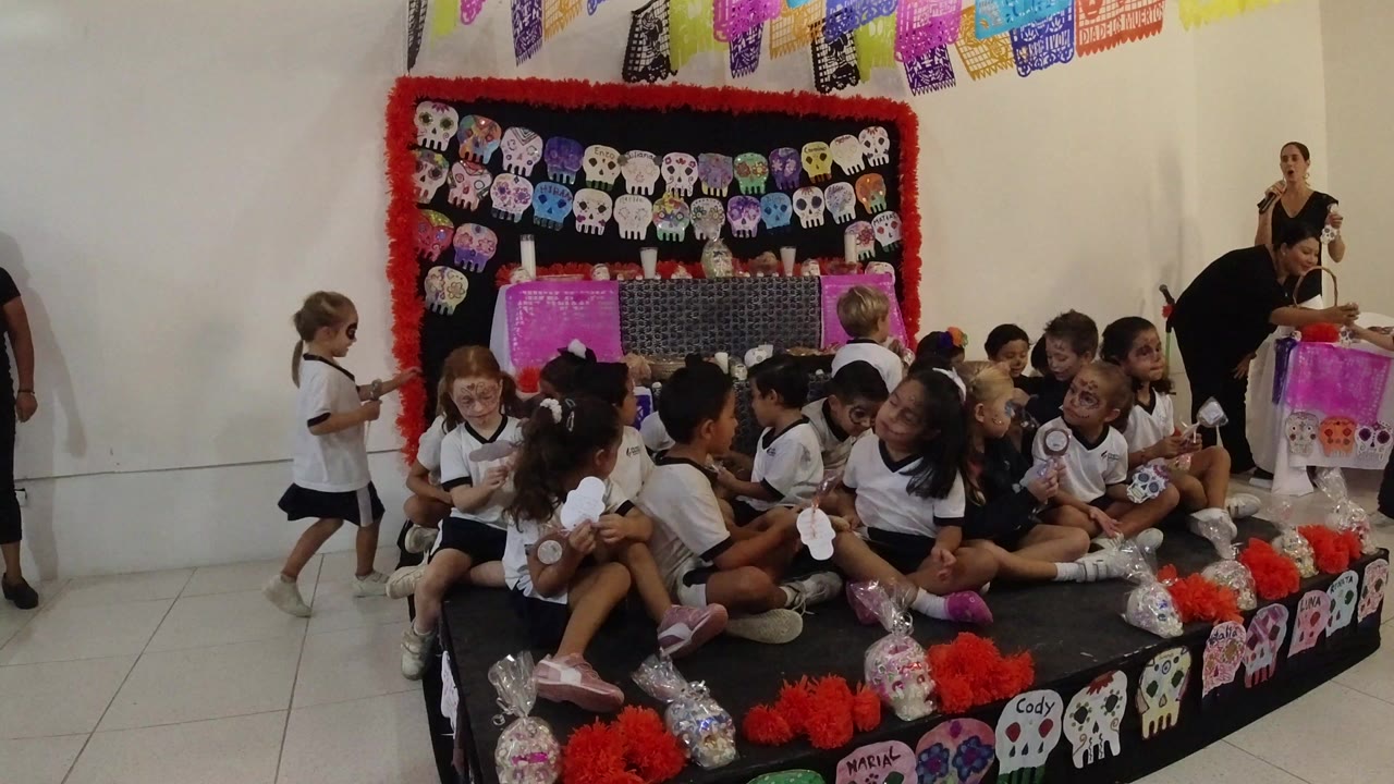 Children in white uniforms sit on a low decorated stage in Los Cabos, Mexico, with skull artwork, red trim, and colorful banners behind them.