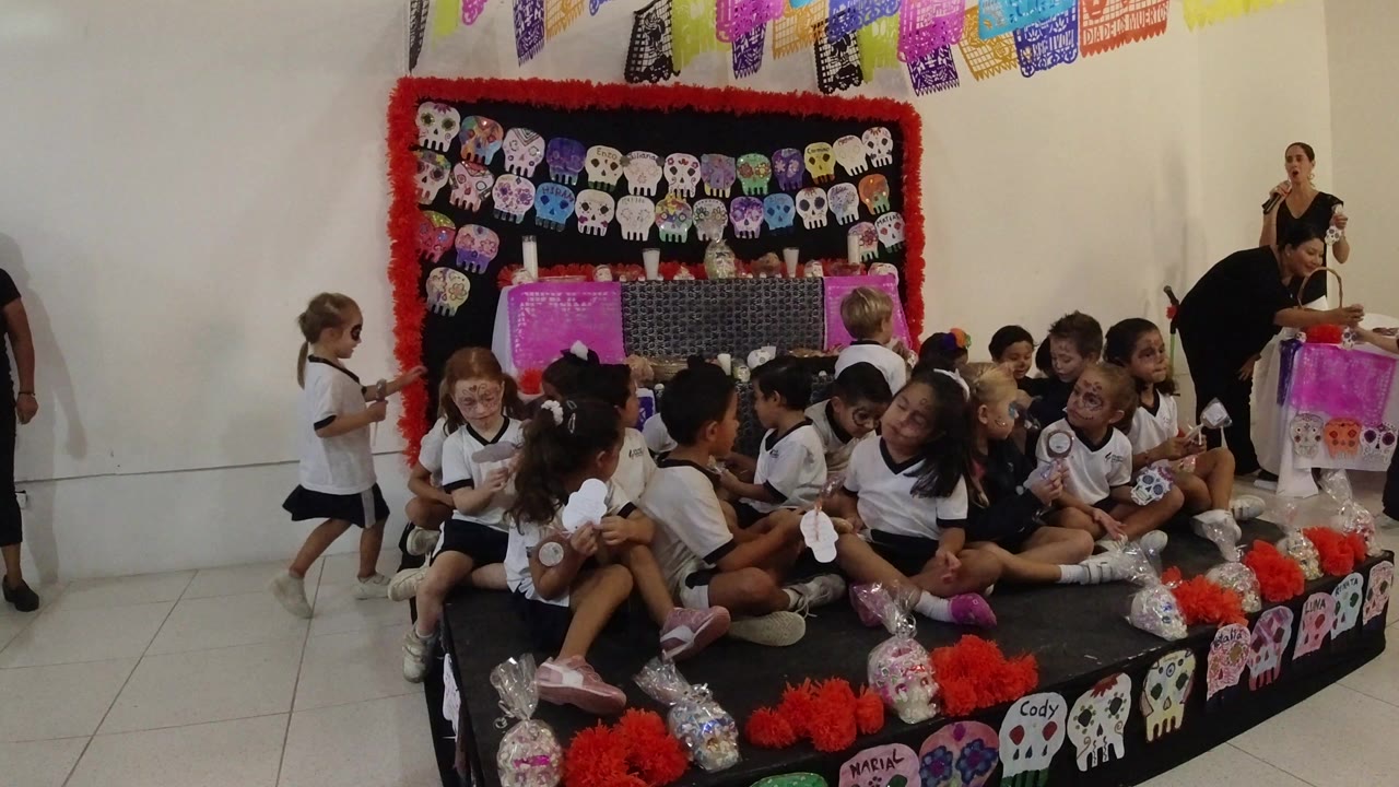 Children sit close together on the stage in Los Cabos, Mexico, while two adults stand near props and decorations at the right side of the frame.