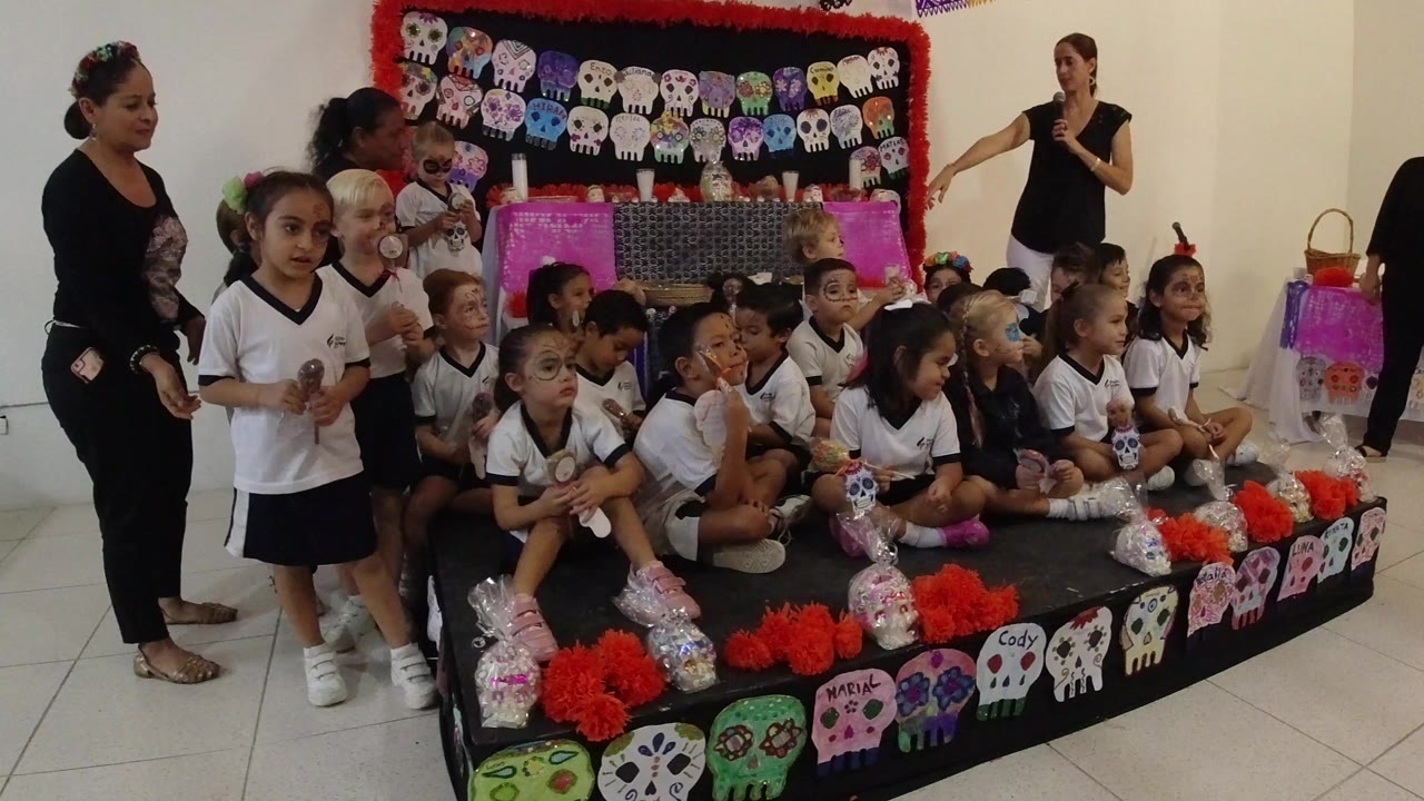 Children in white shirts gather on and beside a low decorated platform in Los Cabos, Mexico, while two adult women stand nearby and colorful skull crafts fill the background.
