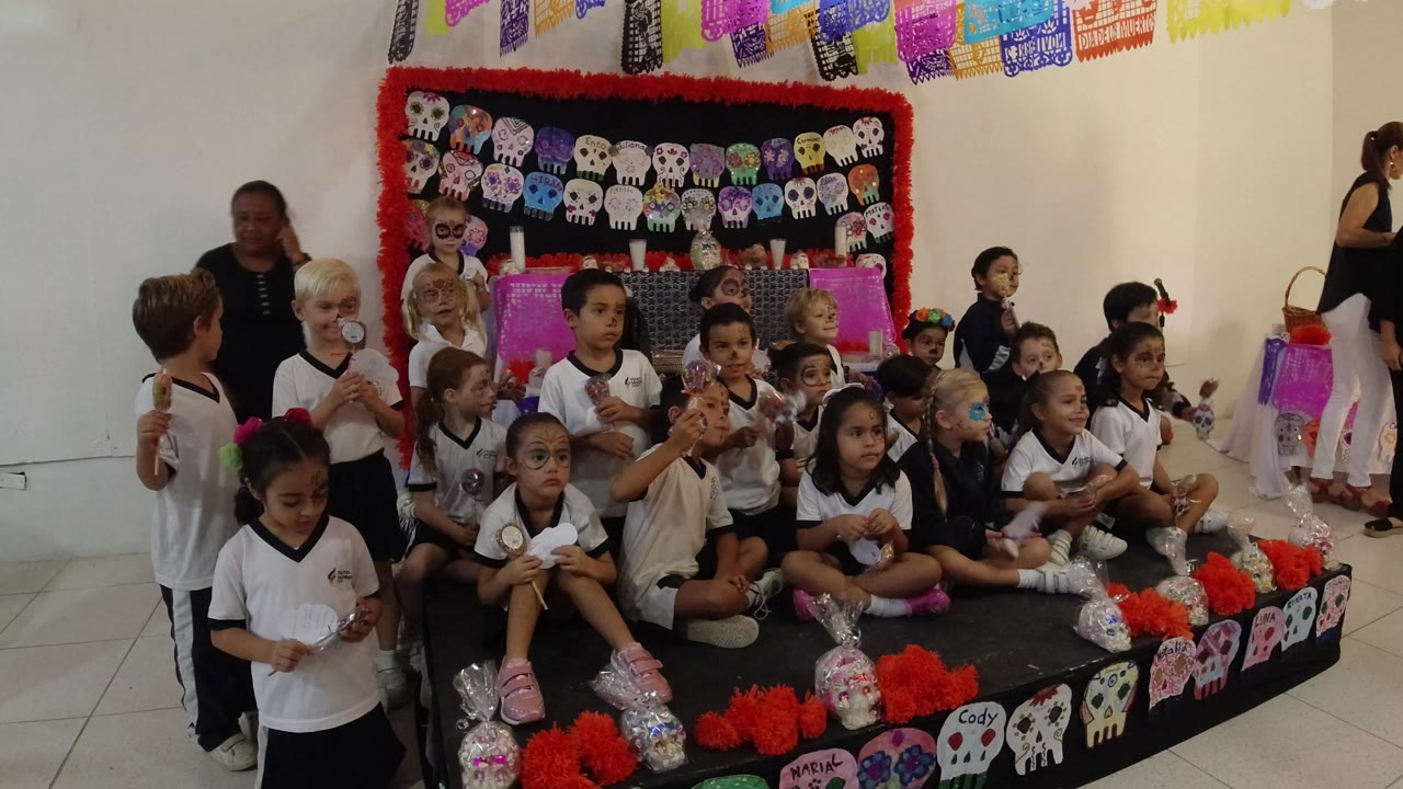 Children in Los Cabos, Mexico sit and stand on a low decorated platform with skull artwork, papel picado banners, and adults nearby at the start of the clip.