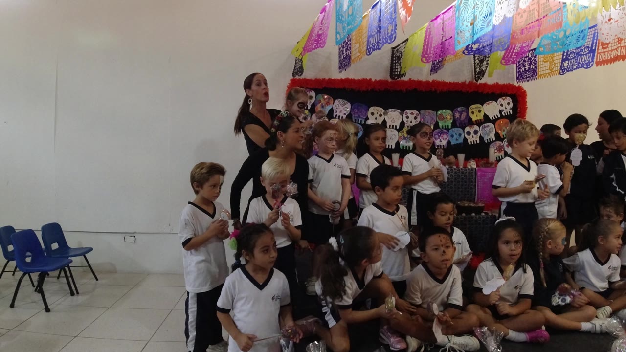 Children in white shirts and adults gather indoors in Los Cabos, Mexico, beneath colorful papel picado and in front of skull-themed wall decorations.