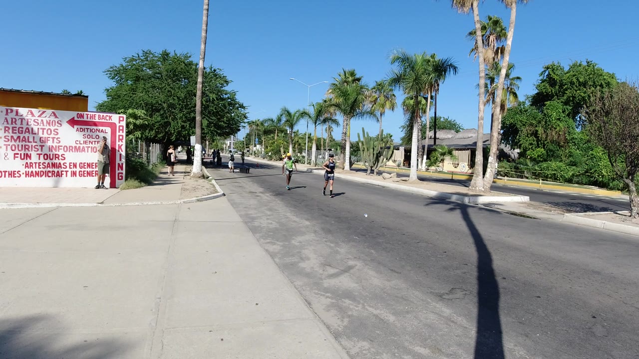 Wide Los Cabos roadside view with a jogger in the lane, another man farther back, palm trees, sidewalk, low buildings, and red plaza signage at the left.