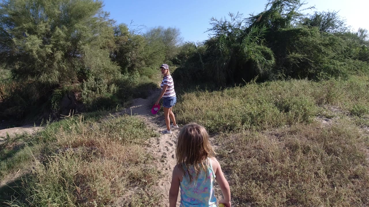 Woman and child on a sandy path surrounded by green brush.