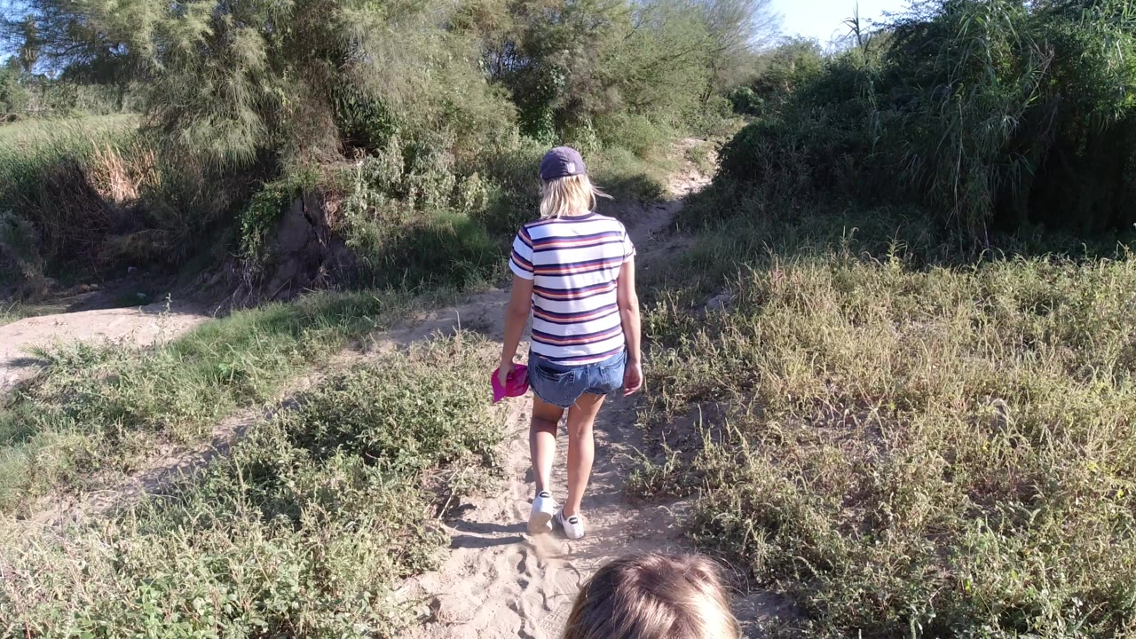 The adult remains centered from behind on the Los Cabos dirt path, holding a bright pink item while bushes and larger trees frame the route ahead.