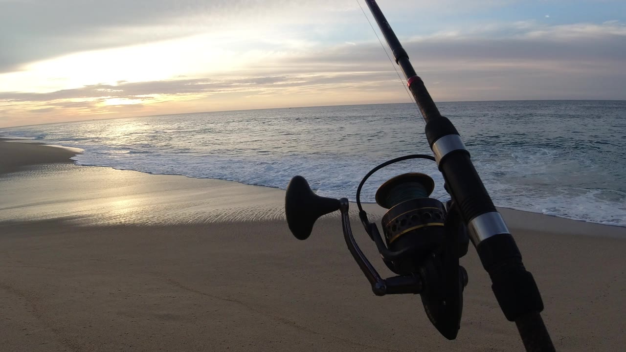 A fishing rod and spinning reel stand in the right foreground on a Los Cabos beach as the sun glows over the ocean and wet sand, with no visible people.