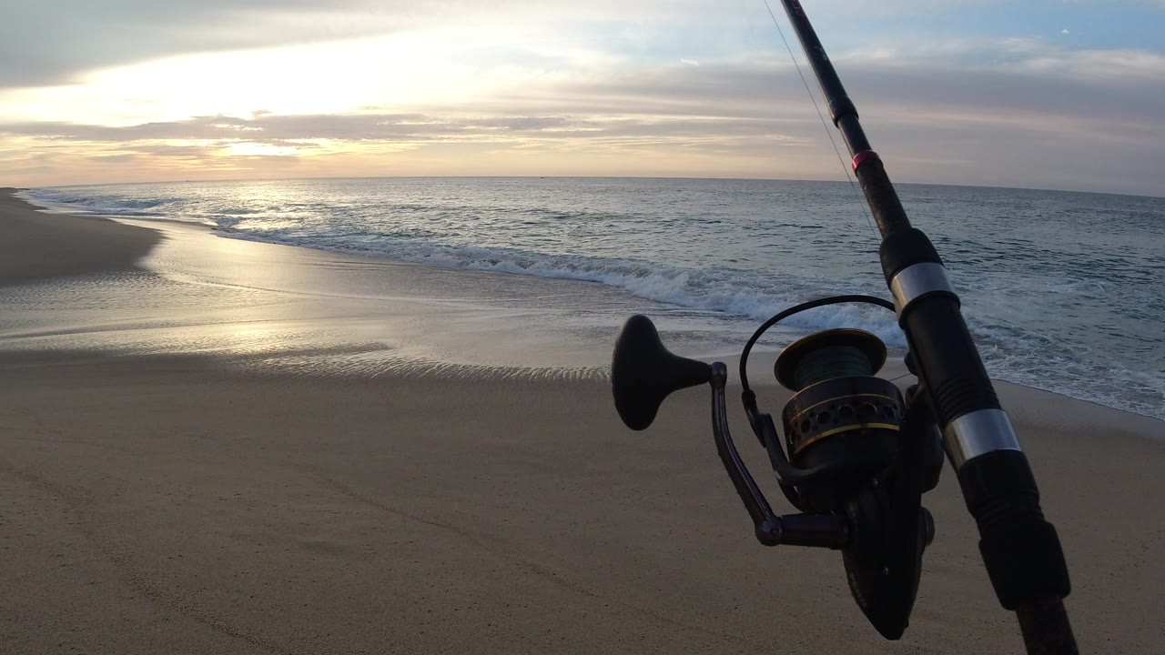 Fishing rod and reel silhouetted beside waves on a sandy beach