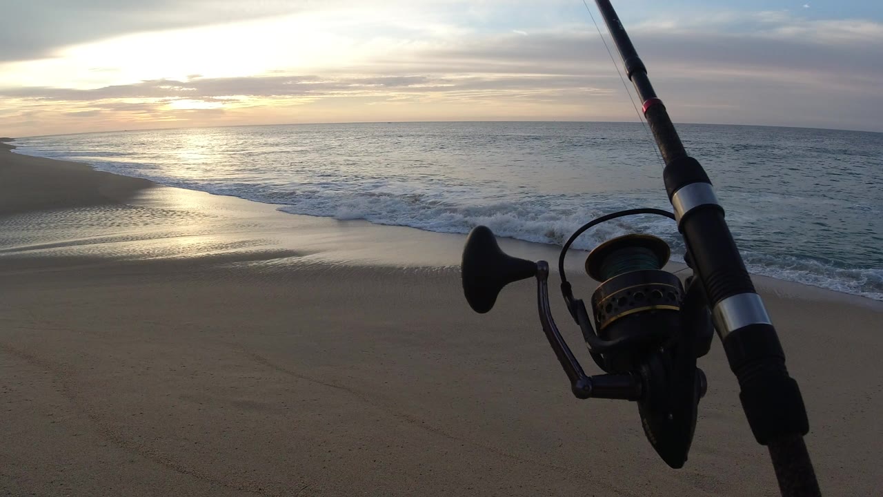 A fishing rod and spinning reel fill the right side of the frame on a Los Cabos beach as sunset light reflects across wet sand and the ocean, with no visible people.