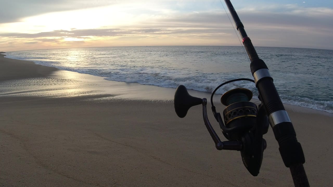 The fishing reel remains near the right foreground while a thin wave line reaches the wet sand under a bright sunset reflection on the Los Cabos shoreline, with no visible people.