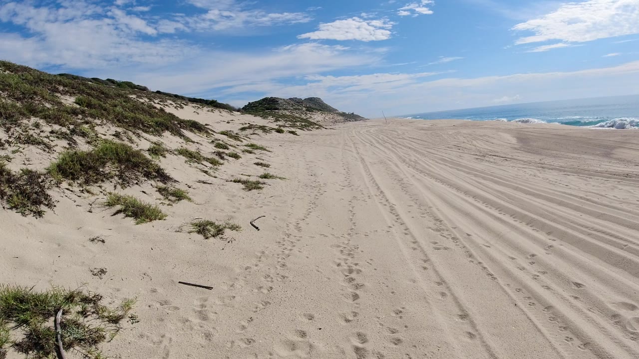 Los Cabos beach view at the start of the clip, with pale sand tracks leading toward the ocean, scrubby dune grass on the left, scattered clouds above, and no visible people.