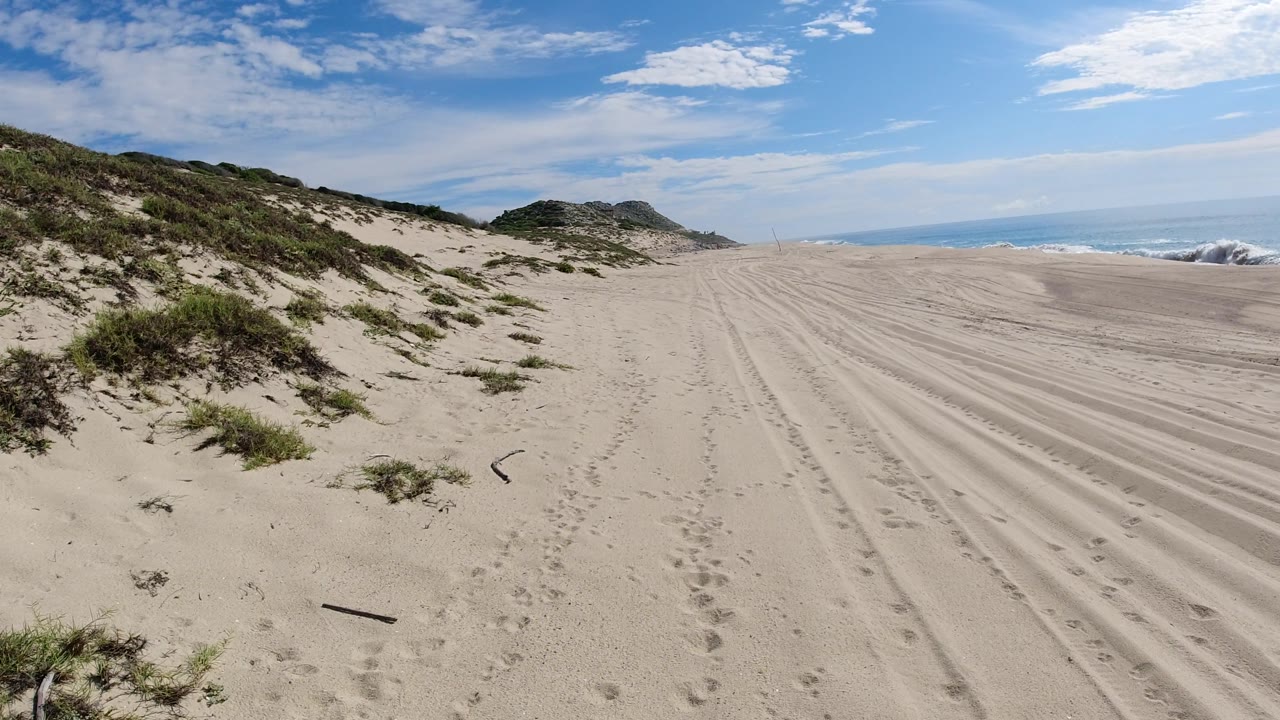 Open sand beside beach grass with ocean surf under a partly cloudy sky.
