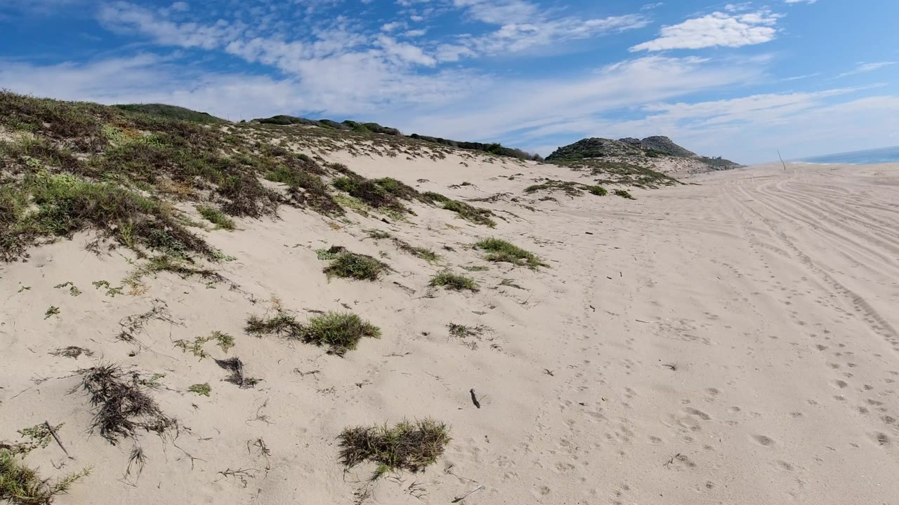 Los Cabos, Mexico sandy dune area with low green brush in the foreground, distant scrub hills, blue sky with scattered clouds, and no visible people.