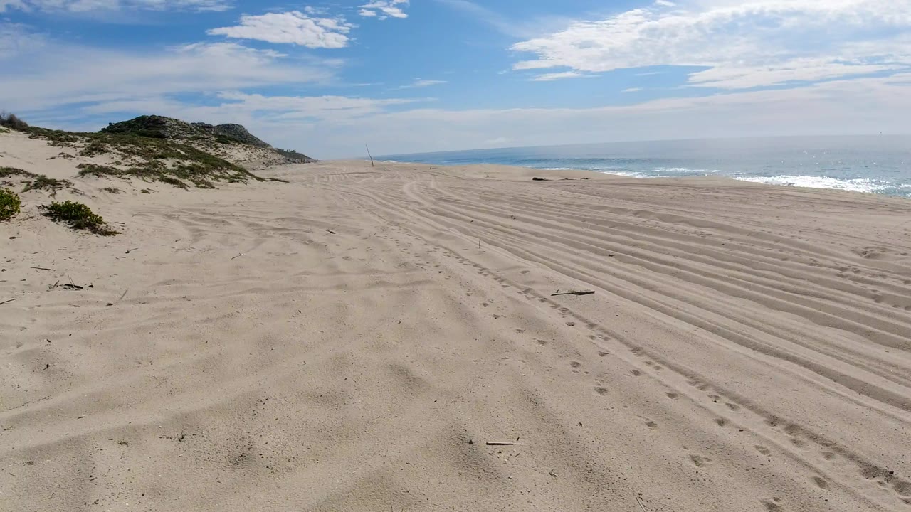 Opening view of a Los Cabos beach with raked-looking tire tracks across pale sand, low dunes at left, blue ocean at right, scattered clouds, and no visible people.