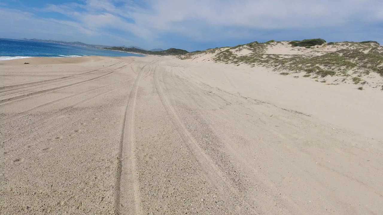 Opening frame of a sandy beach road in Los Cabos, Mexico, with tire tracks leading forward, ocean on the left, scrub-covered dunes on the right, and no visible people.