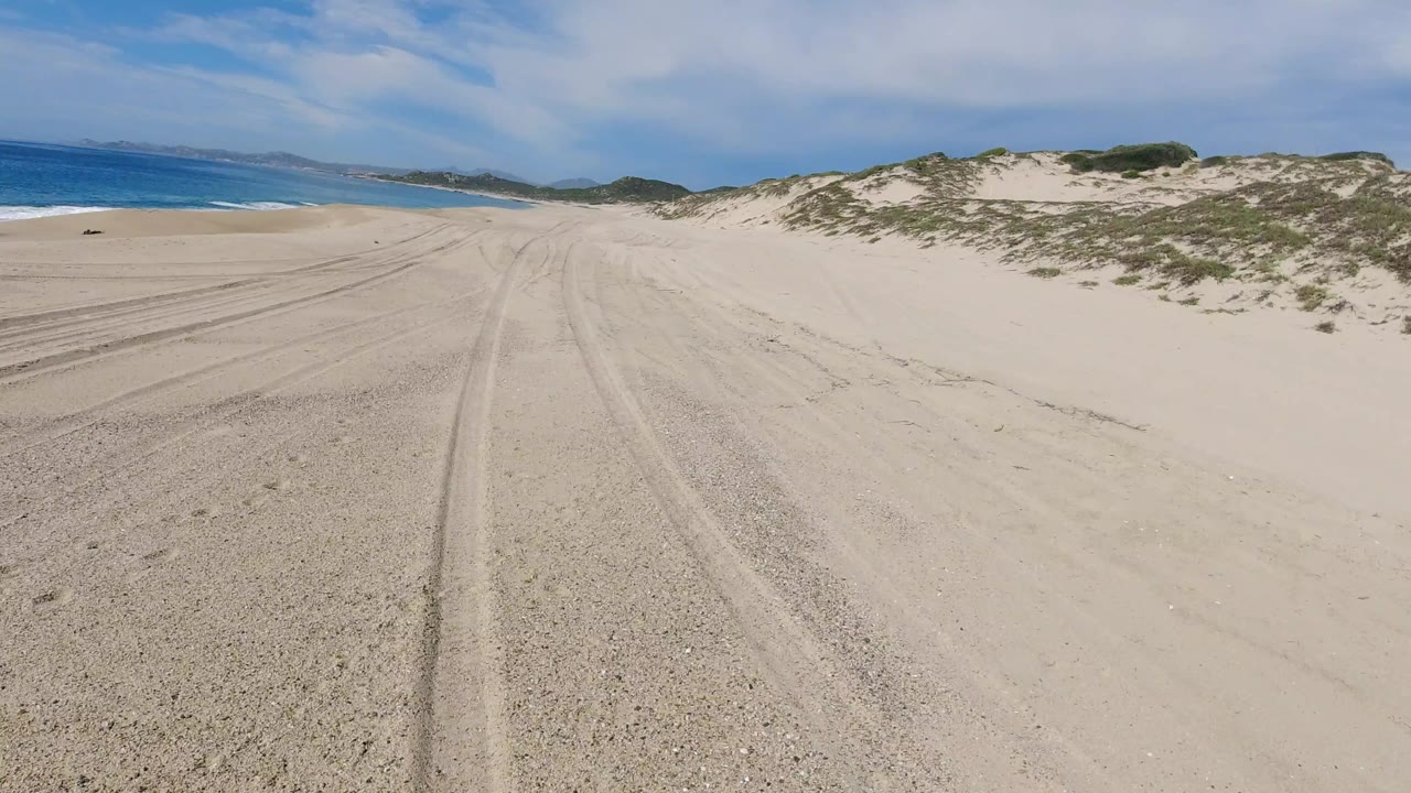 A broad Los Cabos beach-road view shows parallel tire marks across pale sand, the ocean band to the left, dune scrub to the right, and no visible people.