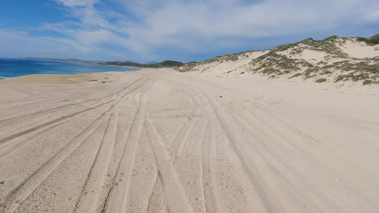 Los Cabos beach sand fills the foreground with multiple tire tracks leading toward distant dunes, ocean on the left, bright clouded sky above, and no visible people.