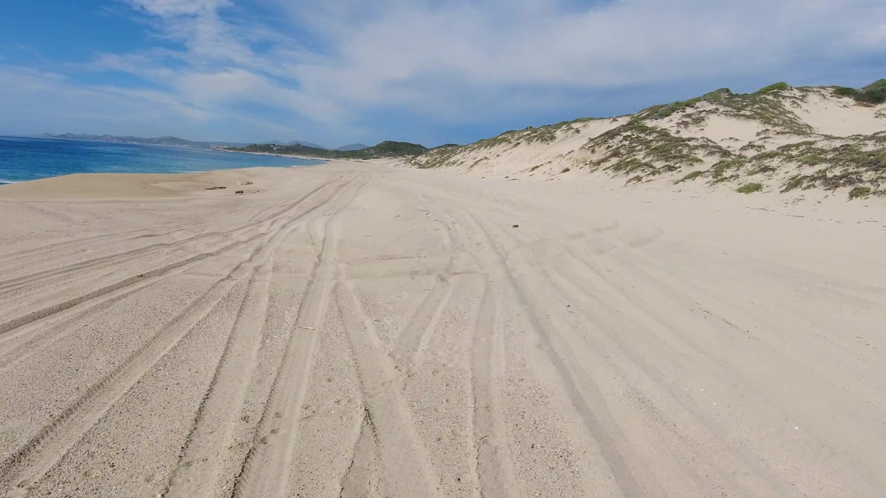 A forward view along pale sand in Los Cabos shows darker tire grooves curving through the beach, blue ocean to the left, dune slope to the right, and no visible people.