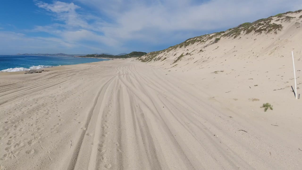 Wide Los Cabos beach view with pale tire tracks in the sand, dunes rising on the right, blue ocean on the left, distant hills ahead, and no visible people.
